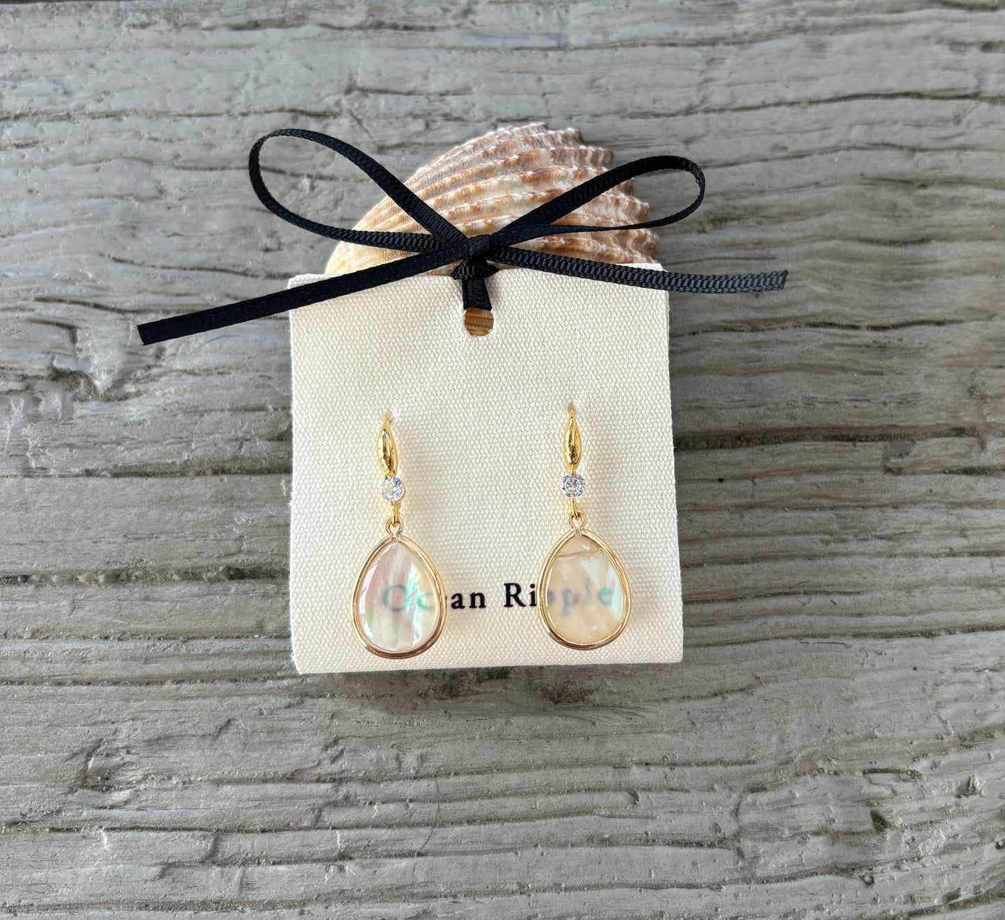 Gold earrings on a branded card with a shell and black ribbon on a textured stone background