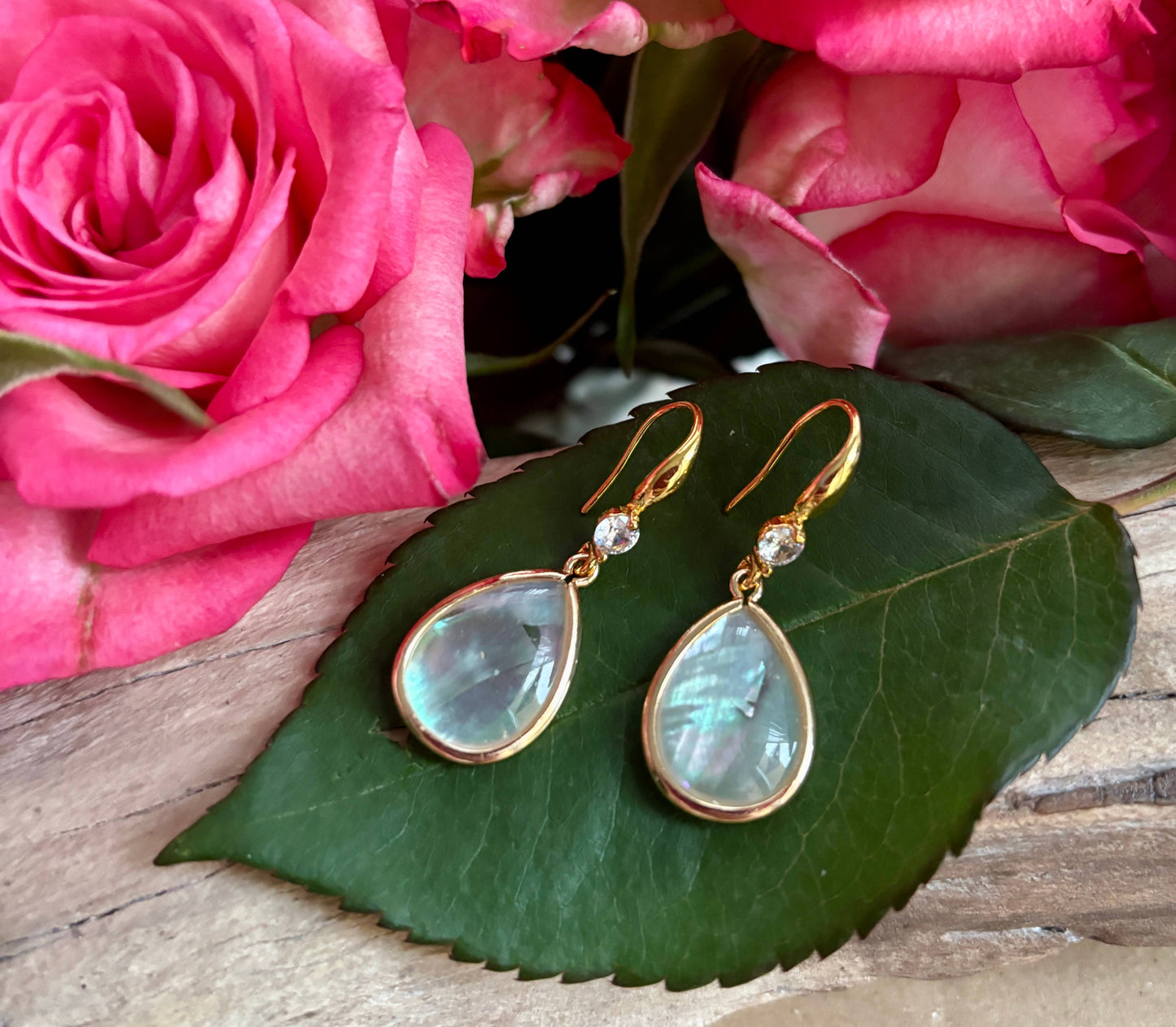 Teardrop-shaped earrings with gold accents on a green leaf with pink roses in the background
