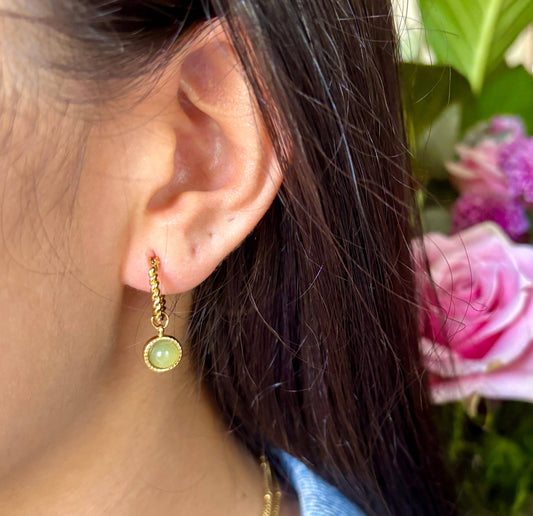 Close-up of an ear wearing a gold earring with a green gemstone, with flowers in the background.