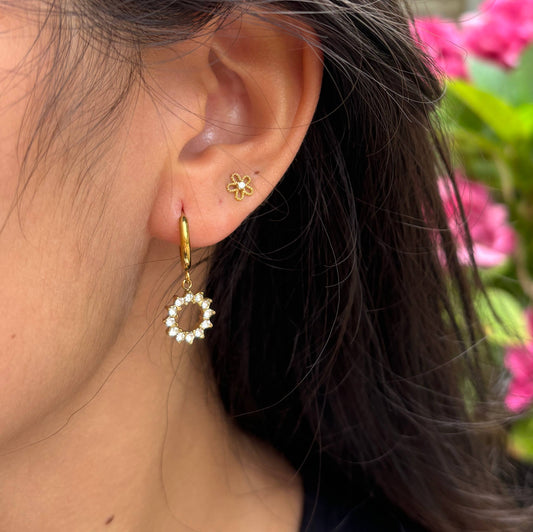 Close-up of a person wearing gold earrings with floral designs, set against a blurred natural background.