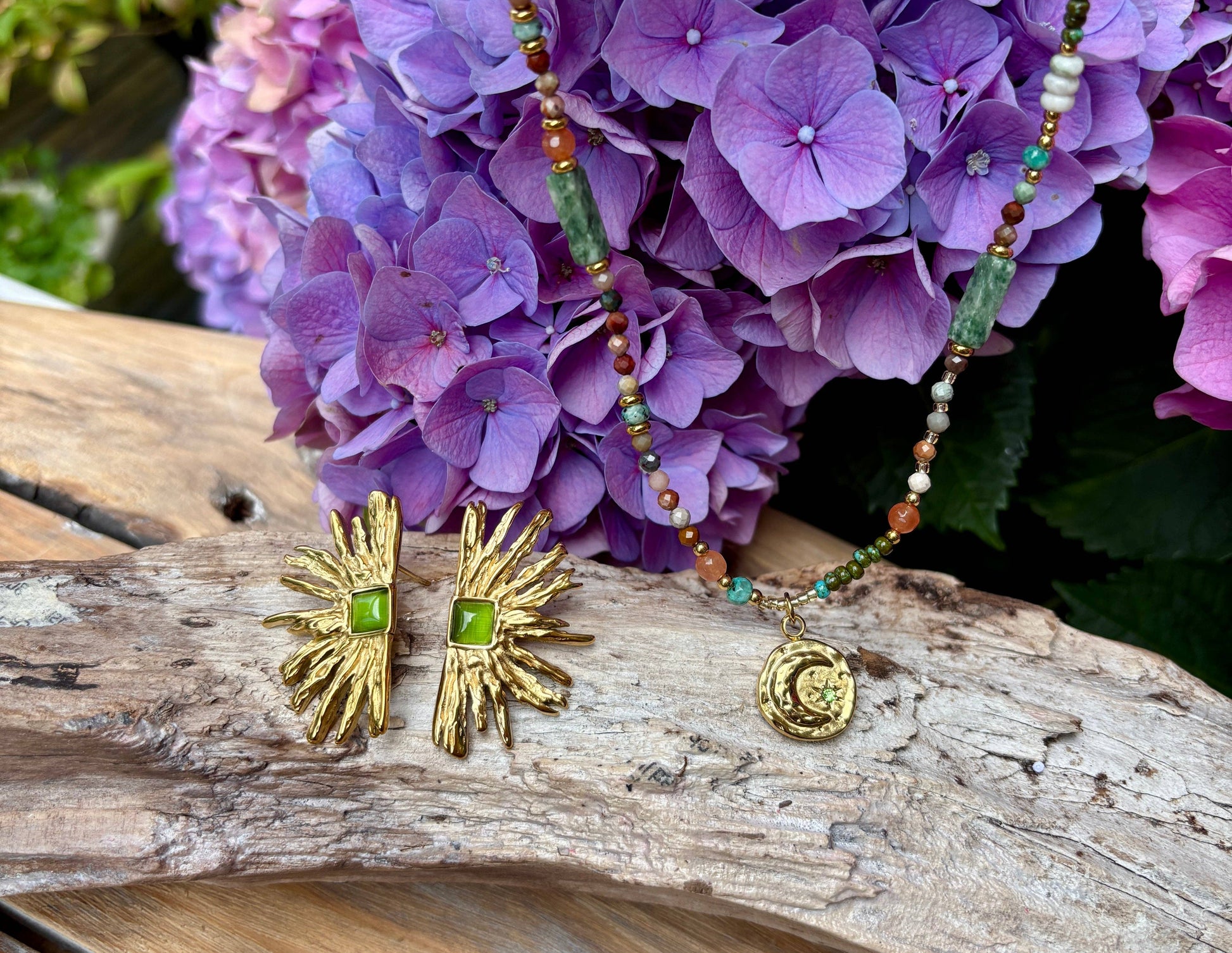 Gold earrings with green stones and a beaded necklace with a pendant on a wooden surface with purple flowers in the background.