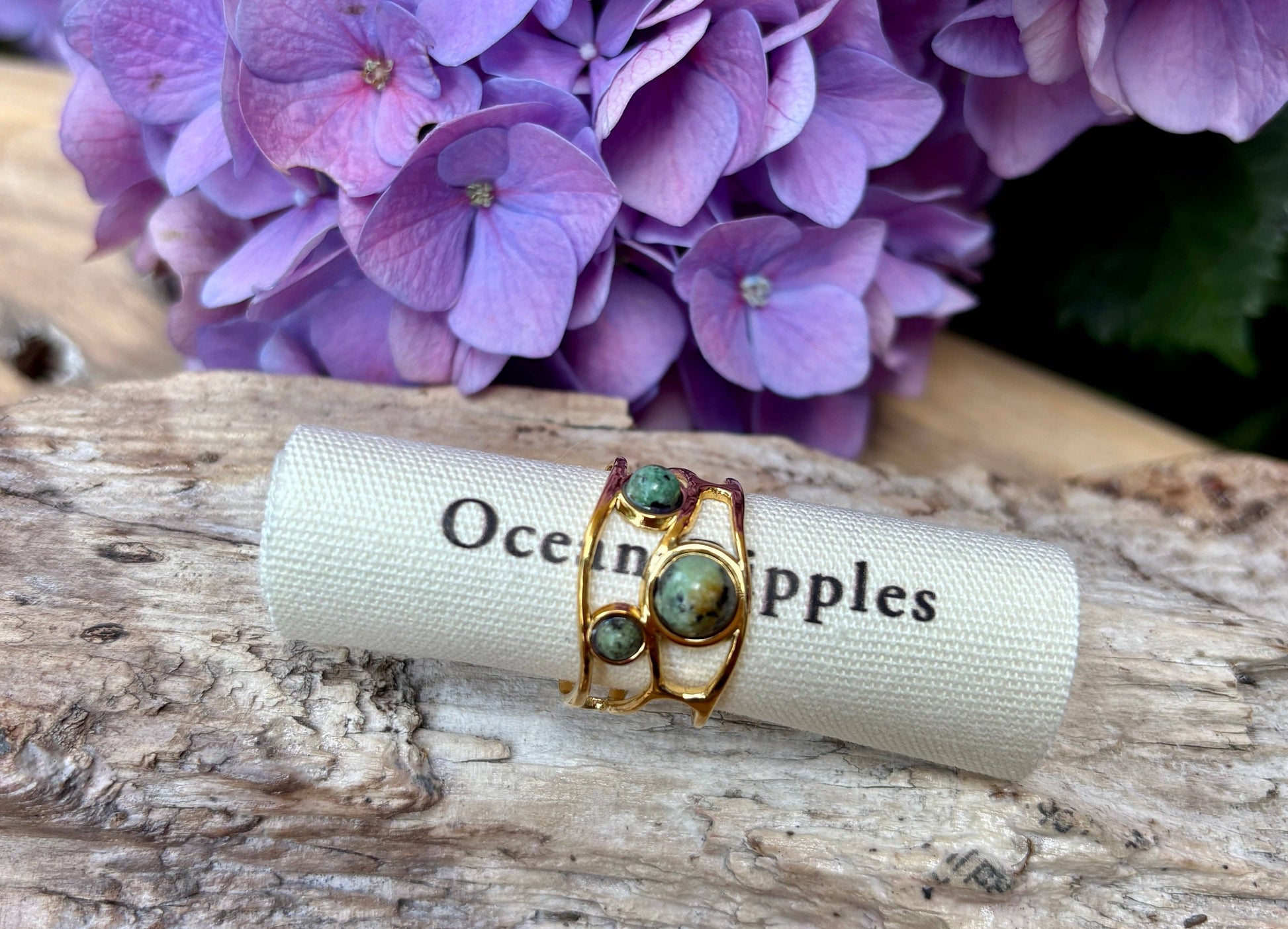 Gold ring with green stones on a piece of paper labeled 'Ocean Apples' with purple flowers in the background.