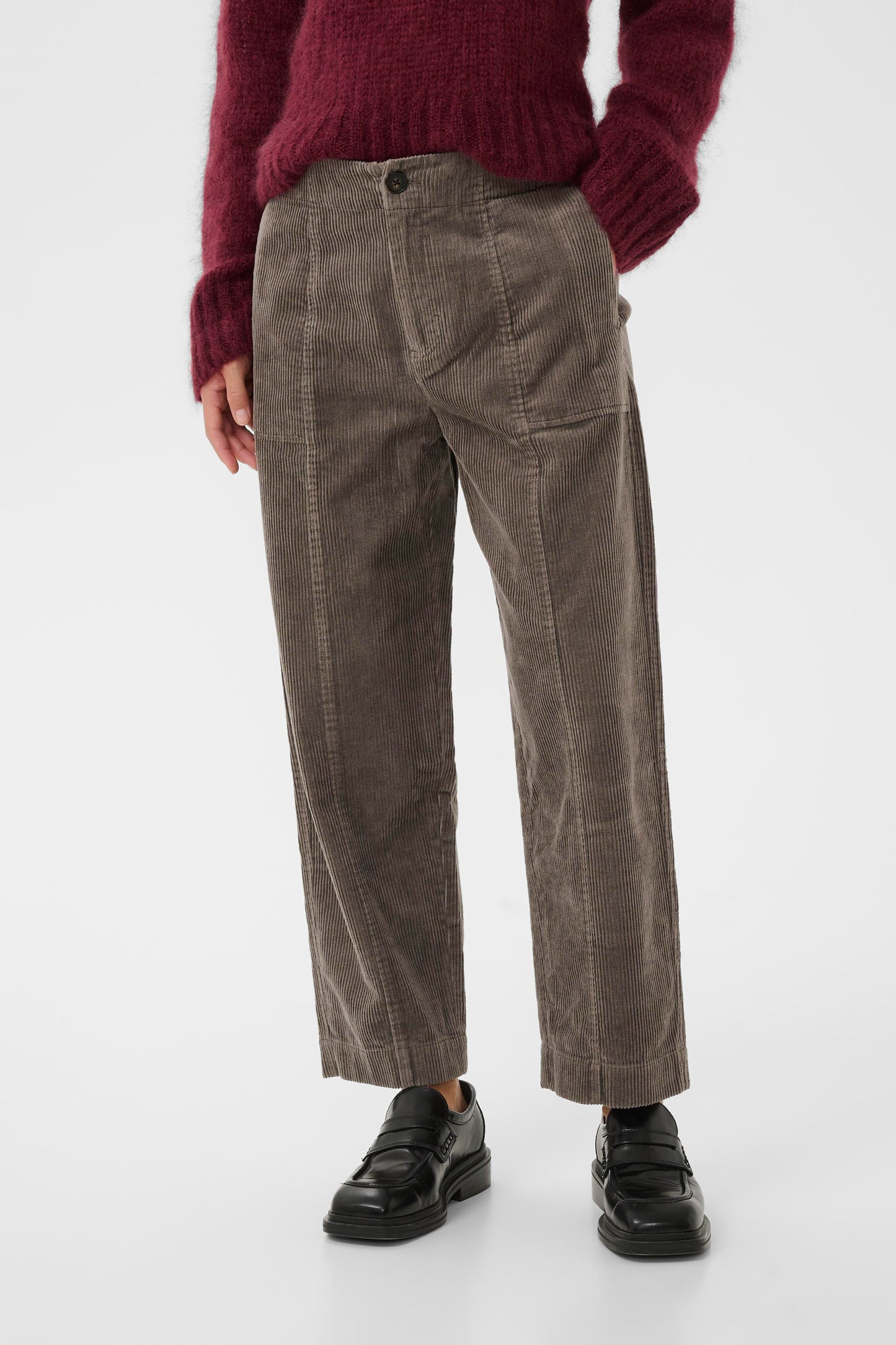 Woman wearing a burgundy sweater and the falcon brown  part two netta cord pants, holding a green tote bag on a white background