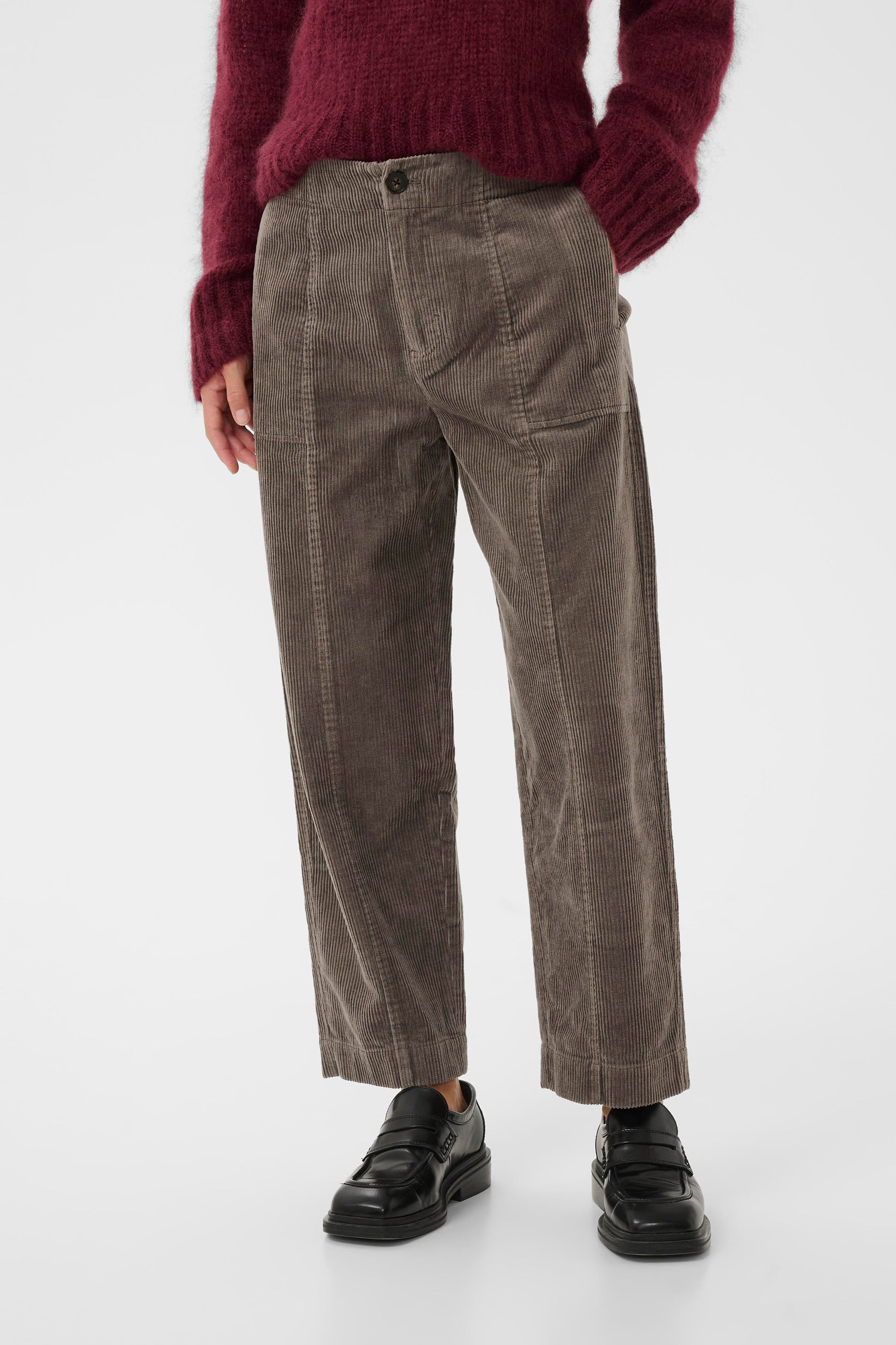 Woman wearing a burgundy sweater and the falcon brown  part two netta cord pants, holding a green tote bag on a white background