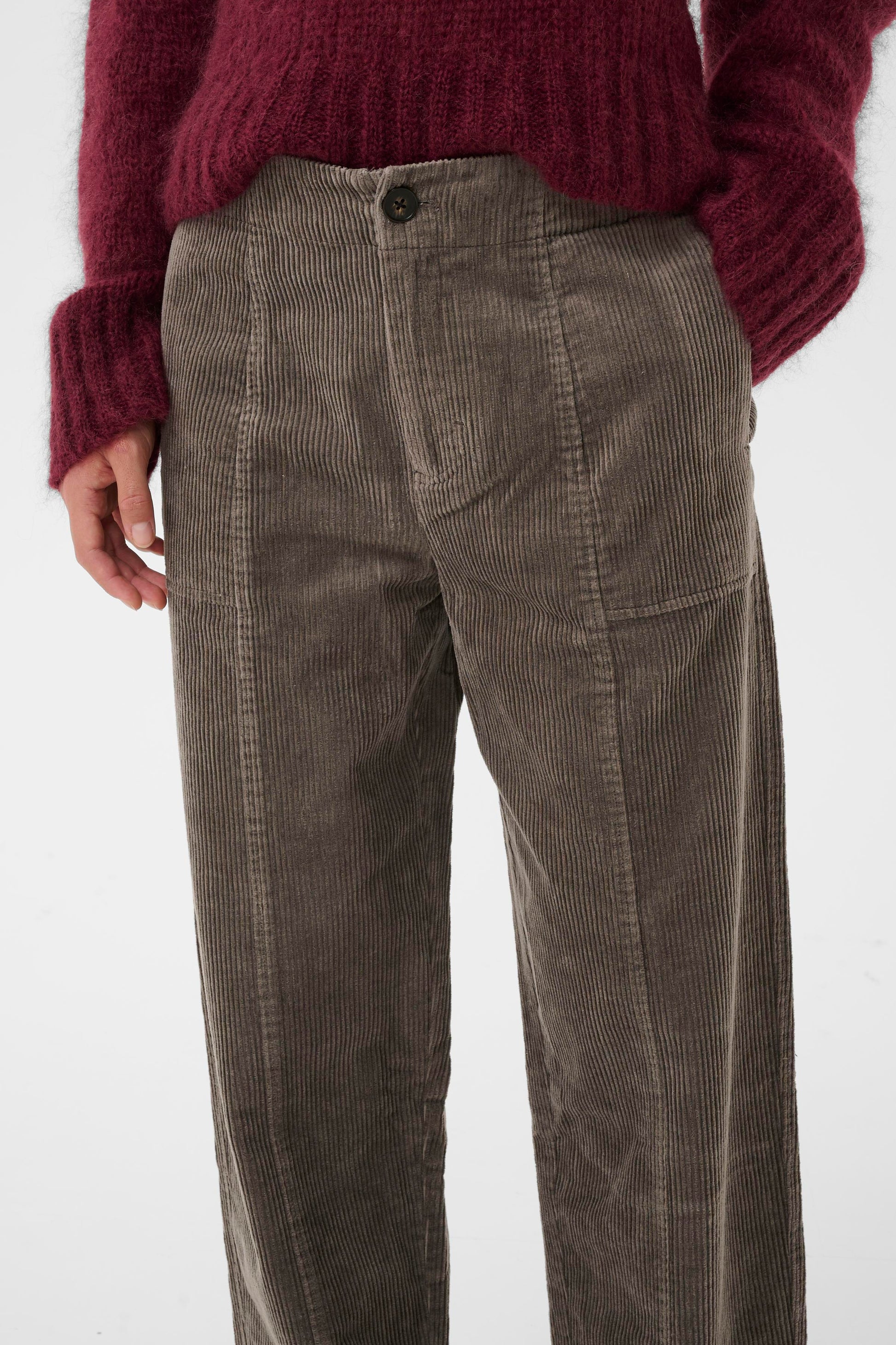 Woman wearing a burgundy sweater and the falcon brown  part two netta cord pants, holding a green tote bag on a white background