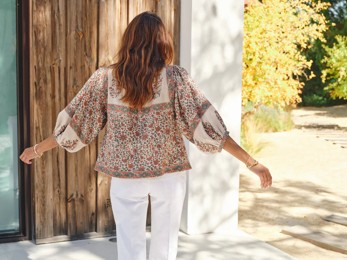 Woman in a della print floral jacket and white pants standing in a sunlit outdoor setting.