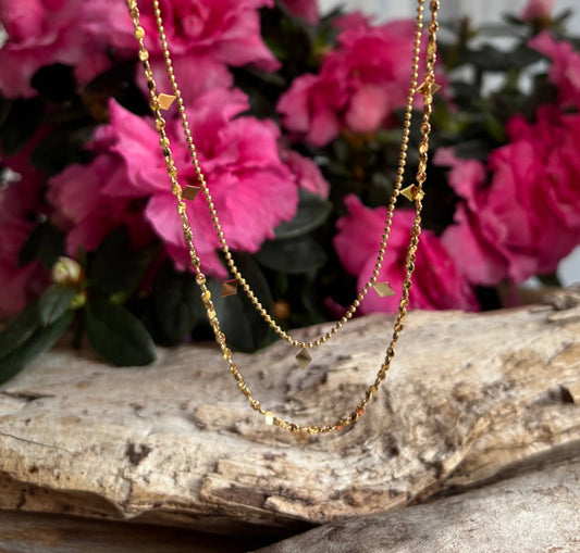 double layer gold necklace with small detailing on.  Displayed on a wooden plinth and pink flowers