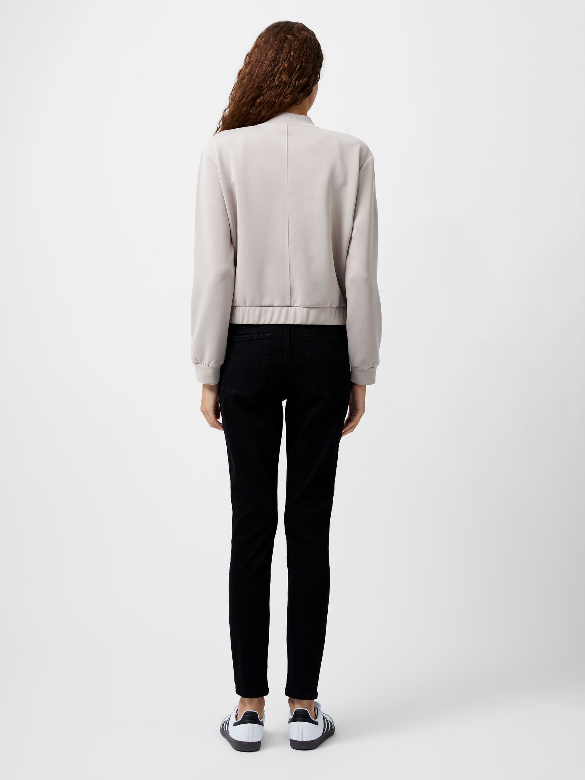 Woman wearing a cropped khaki colour zip up bomber jacket.  She is wearing a pair of black jeans too.  View from the back