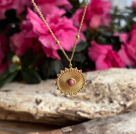 Gold necklace with a pink pendant on a stone surface with pink flowers in the background