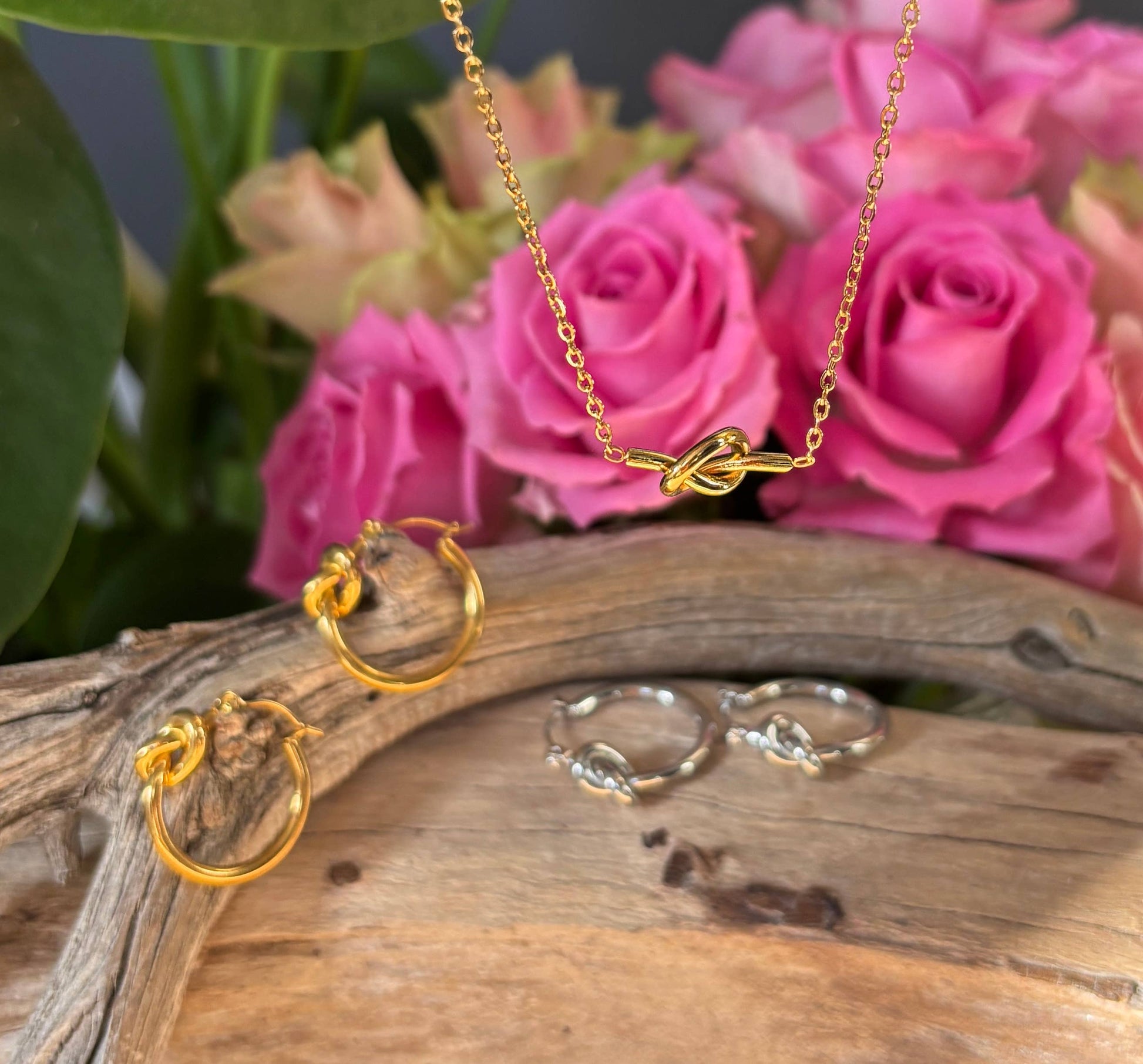 Gold jewelry set including necklace, earrings, and ring on a wooden surface with pink roses in the background.