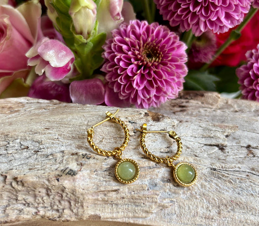 Gold hoop earrings with green stones on a wooden surface with pink flowers in the background