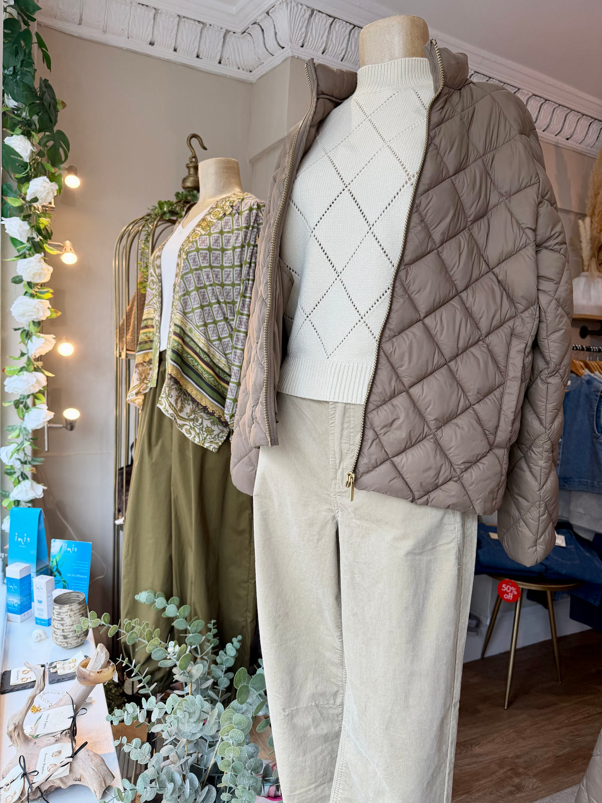 A mannequin in a boutique displays a three-piece outfit consisting of a taupe quilted puffer jacket, a cream argyle sweater, and cream corduroy trousers. Other clothing items are visible in the background of the shop.