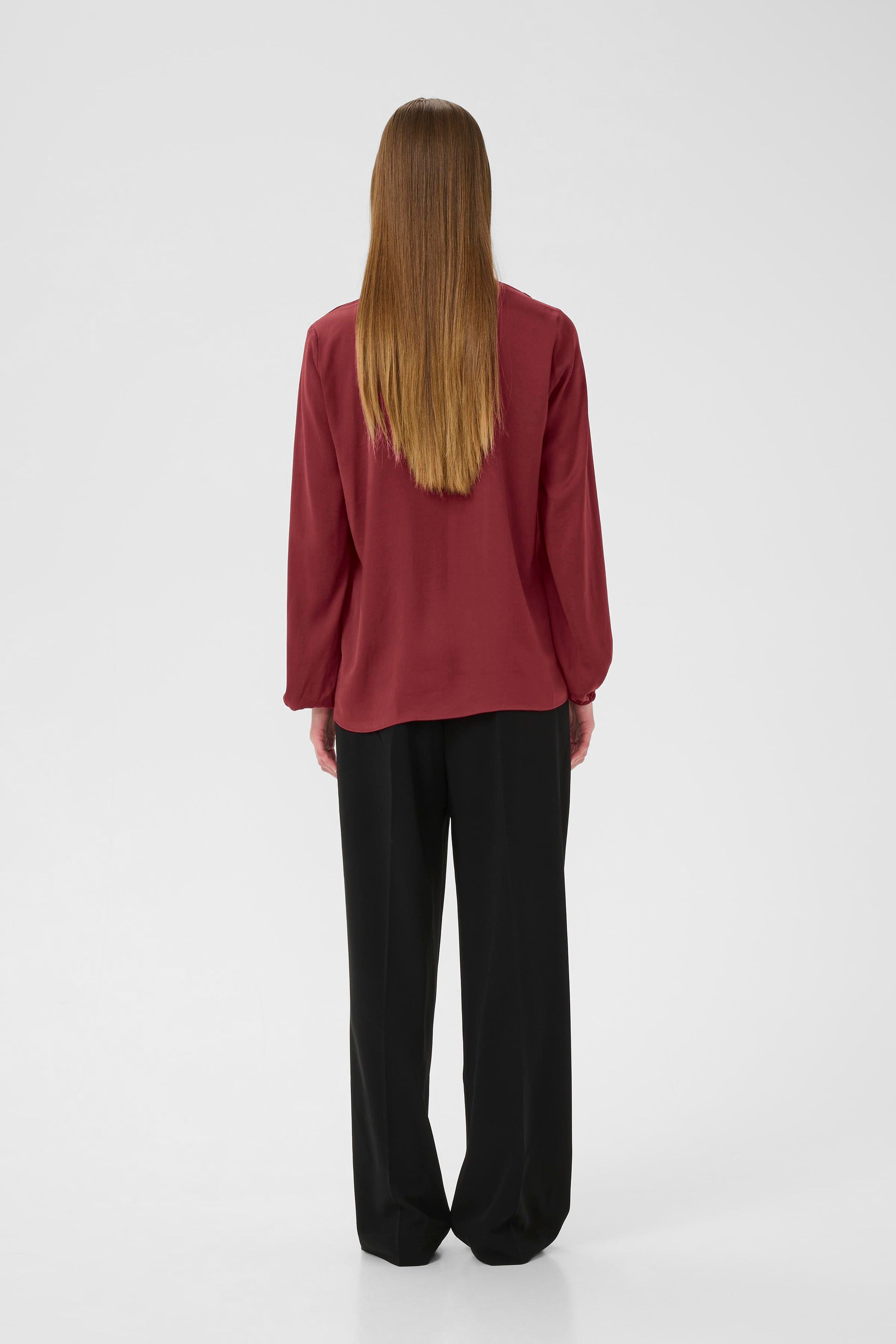 Woman wearing a burgundy Blouse with long sleeves and V Neck on a white background.  View is from the back