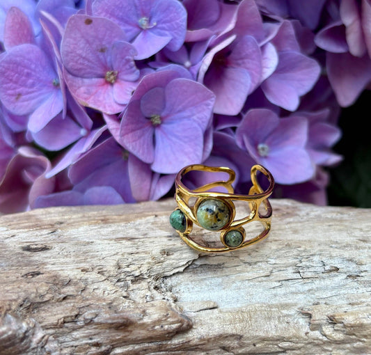 Gold ring with green stones on a wooden surface with purple flowers in the background