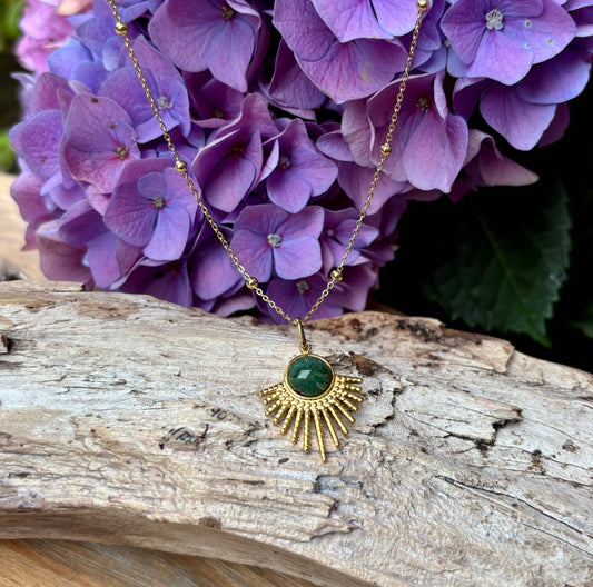 Gold necklace with a green pendant on a wooden surface with purple flowers in the background