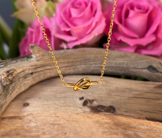 Gold necklace with a knot pendant on a wooden surface with pink roses in the background