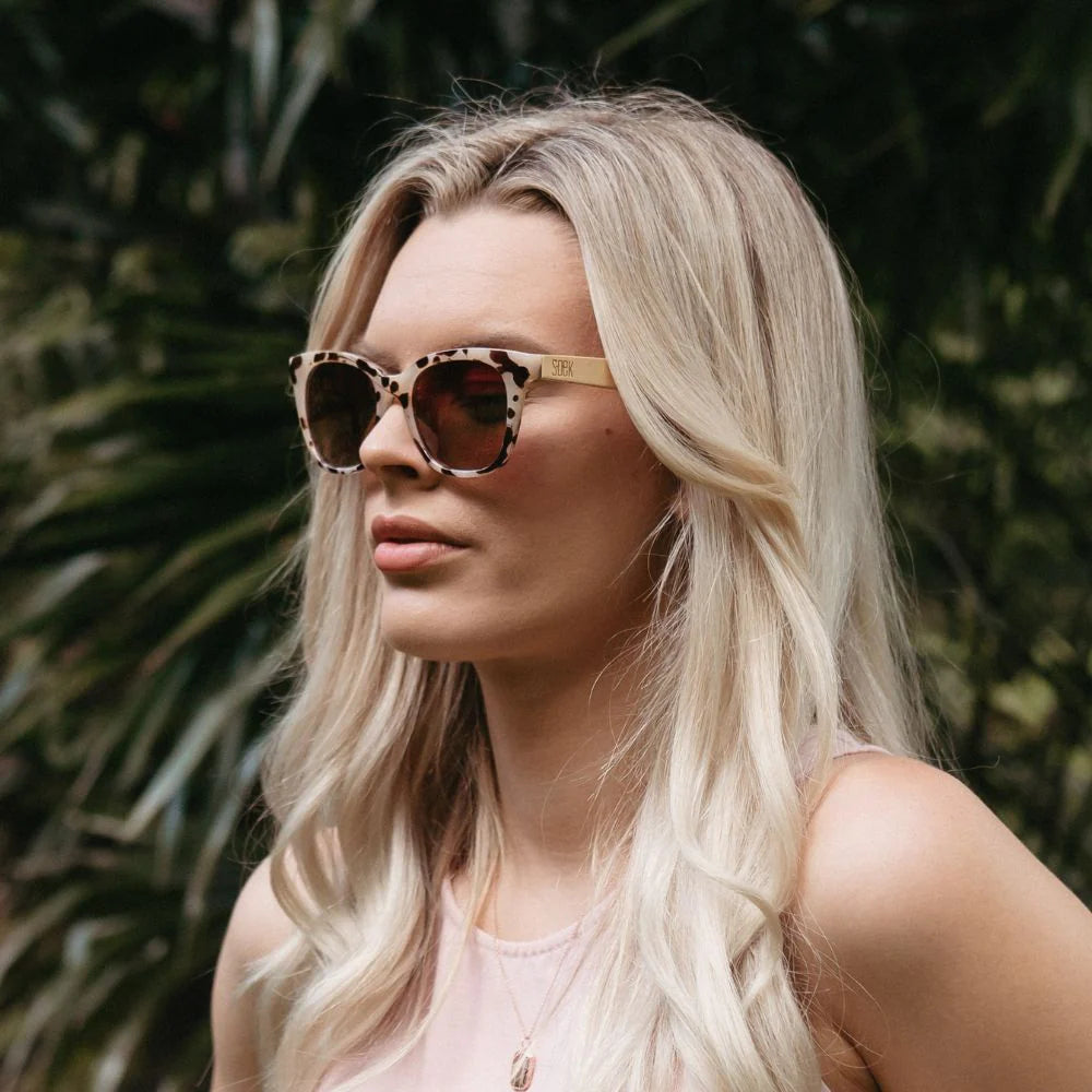 Sunglasses with tortoiseshell frame and brown gradient lenses, accompanied by a wooden arm on a white background.