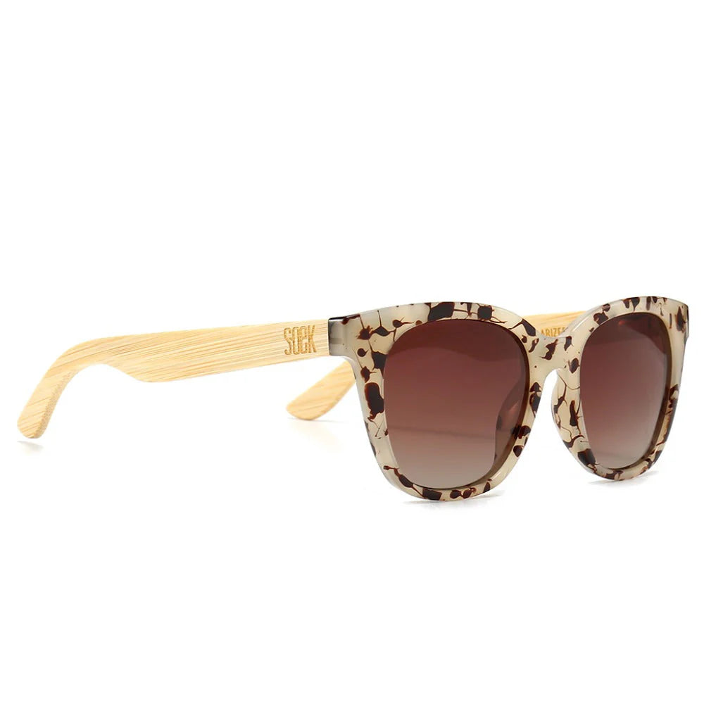 Sunglasses with tortoiseshell frame and brown gradient lenses, accompanied by a wooden arm on a white background.