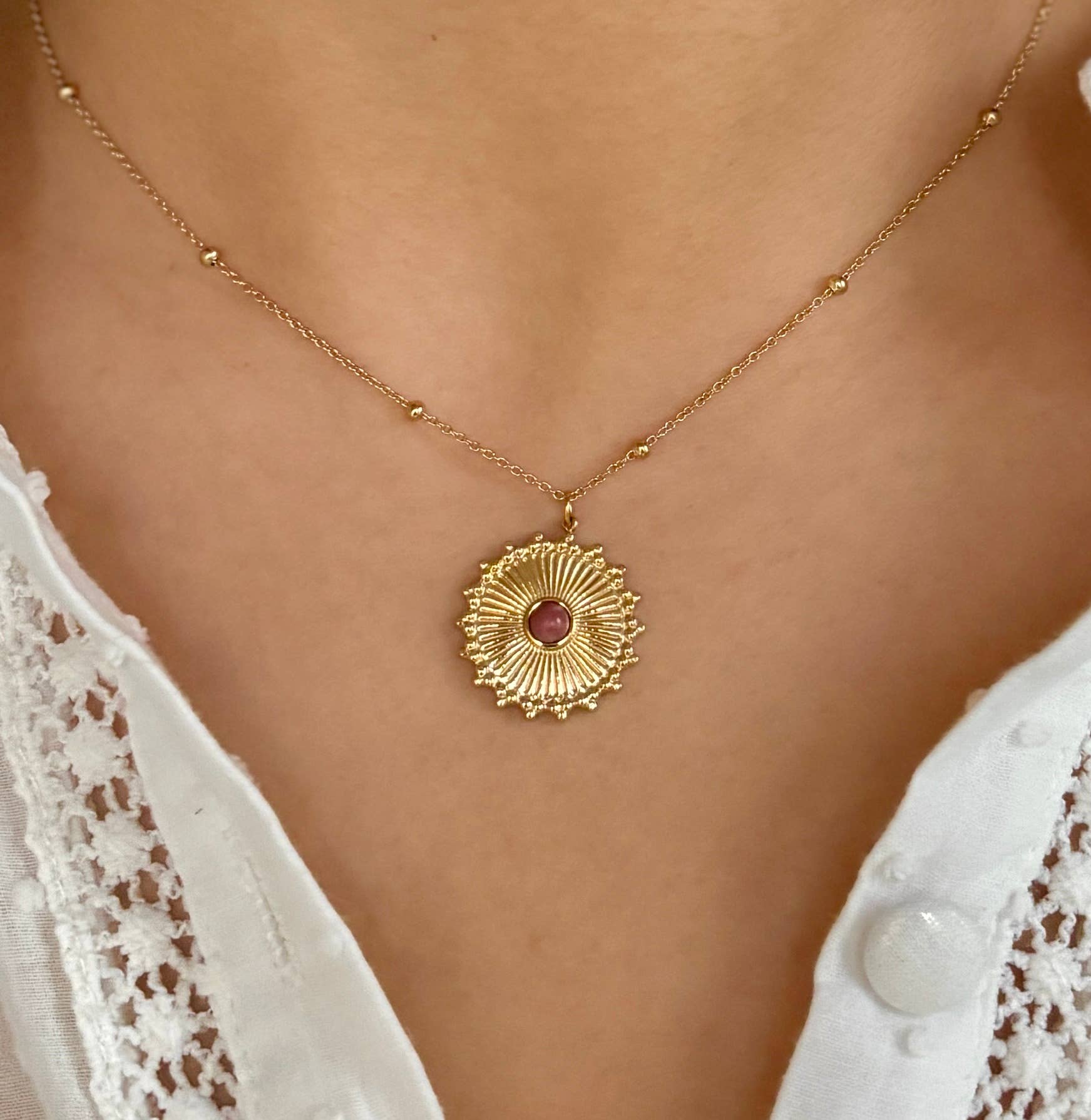 Gold necklace with a pink gemstone pendant on a person wearing a white lace top.