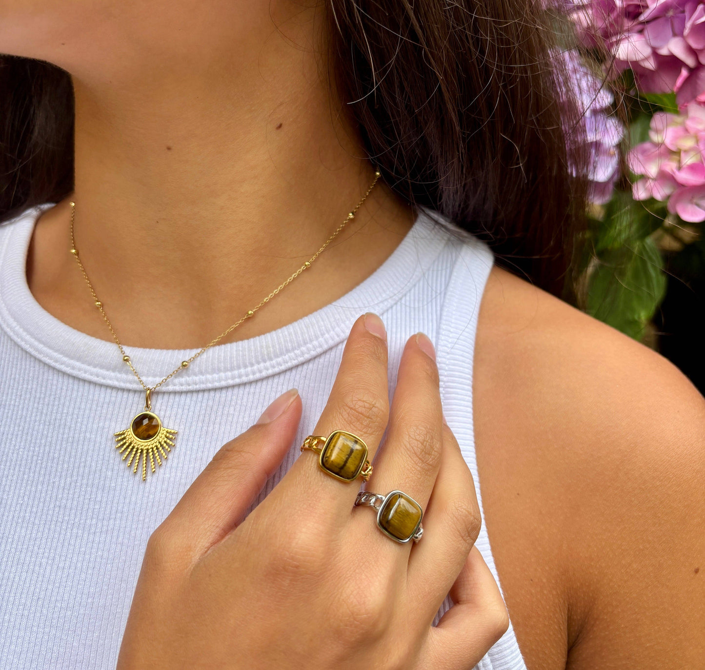 Person wearing a gold necklace and ring with a white top and flowers in the background