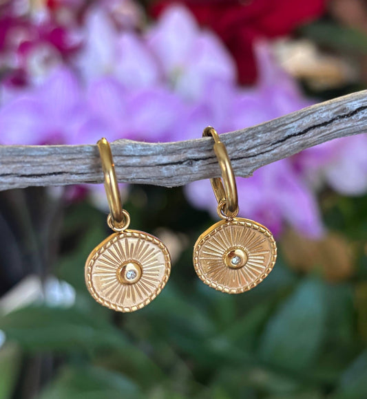 Gold plated earrings with circular designs and cubic zirconia stone hanging on a branch with blurred flowers in the background