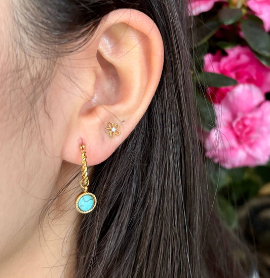 Close-up of an ear wearing a gold earring with a turquoise stone, with pink flowers in the background.