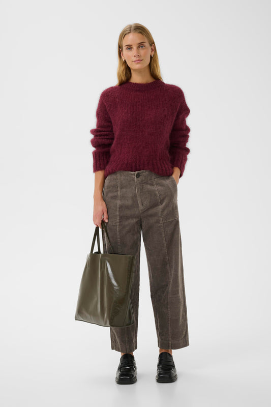 Woman wearing a burgundy sweater and the falcon brown  part two netta cord pants, holding a green tote bag on a white background