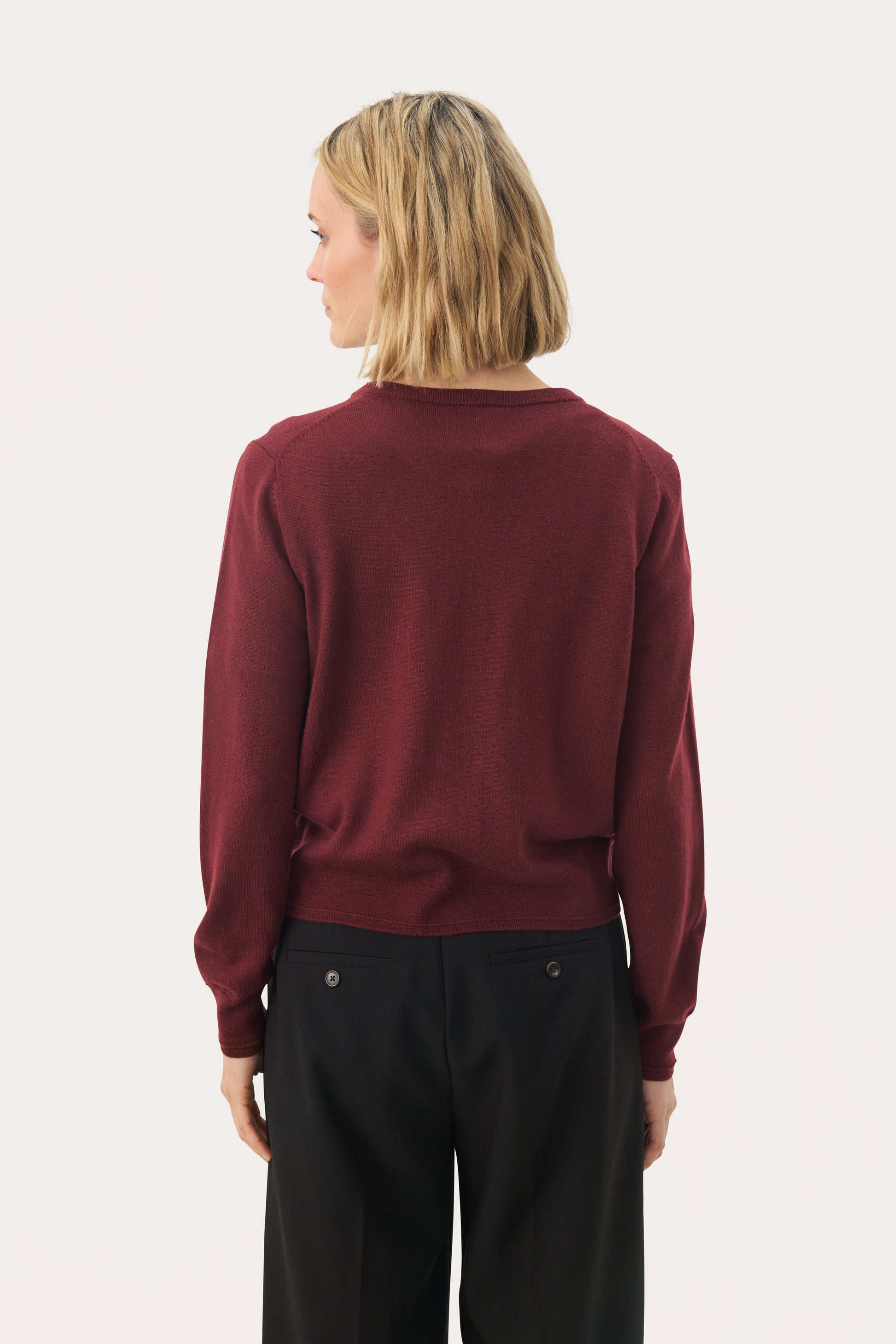 Woman wearing a burgundy sweater and black pants on a white background.  The view is from the back