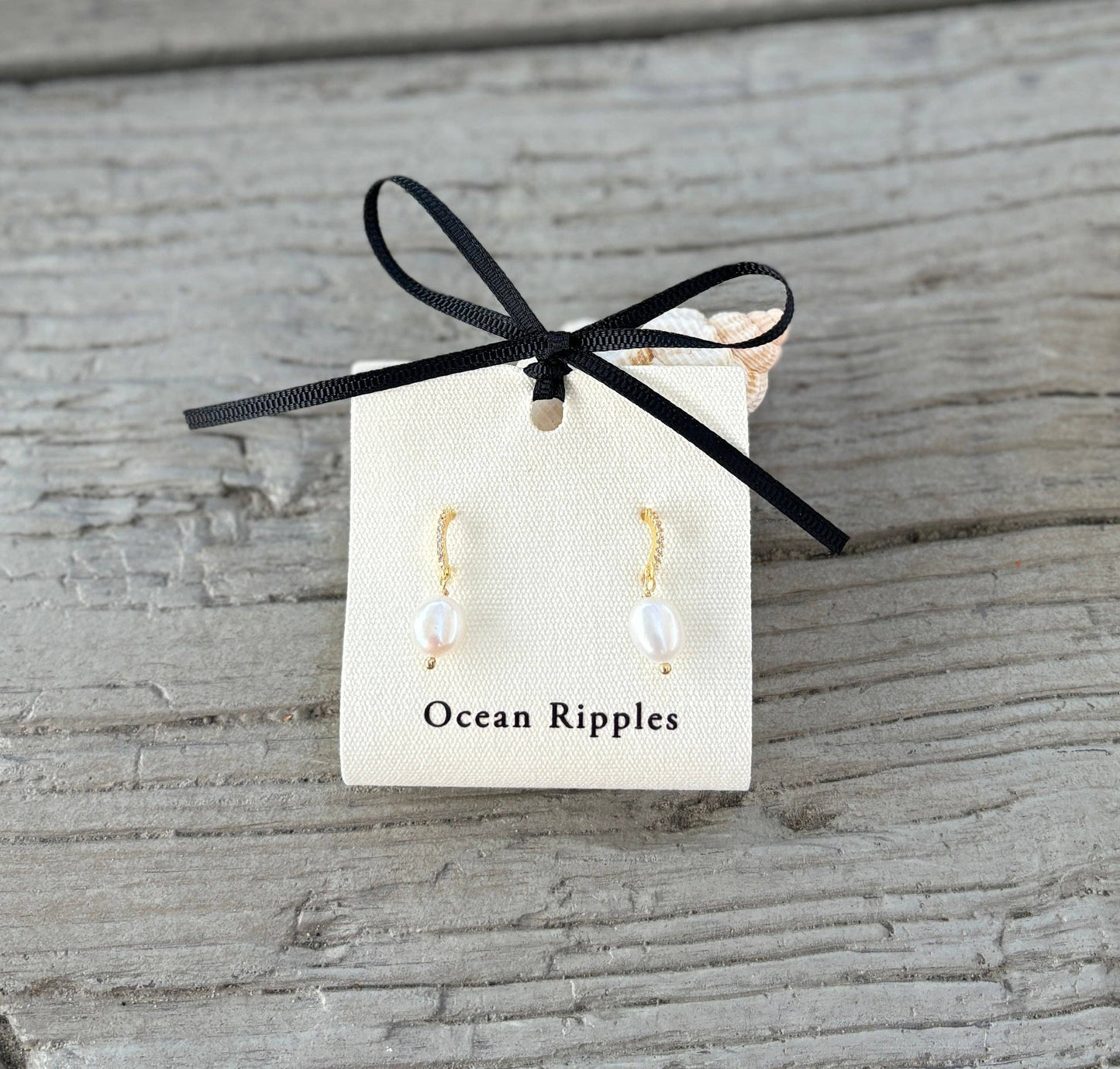 Pearl earrings on a card labeled 'Ocean Ripples' with a black ribbon on a wooden surface.