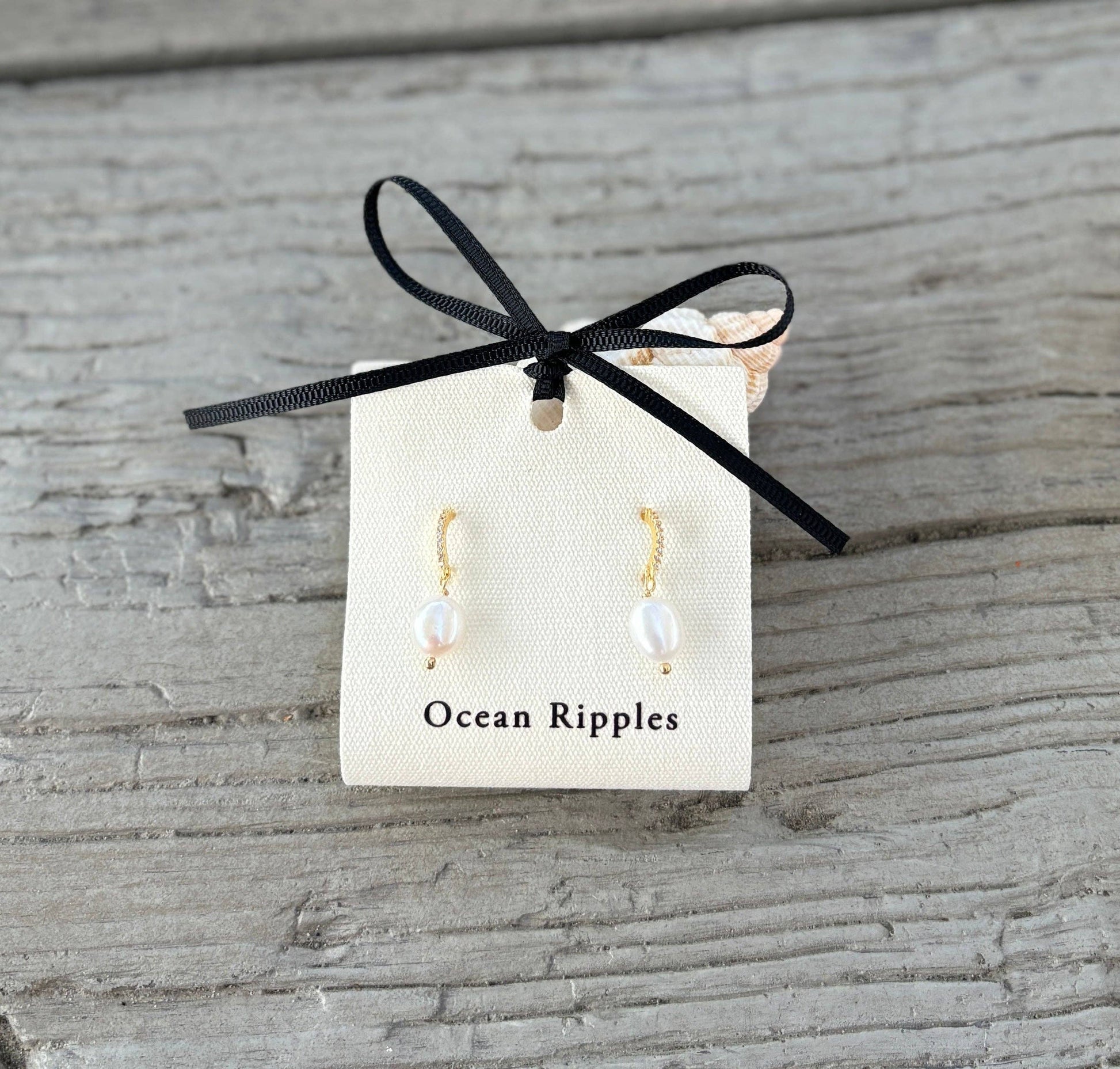 Pearl earrings on a card labeled 'Ocean Ripples' with a black ribbon on a wooden surface.