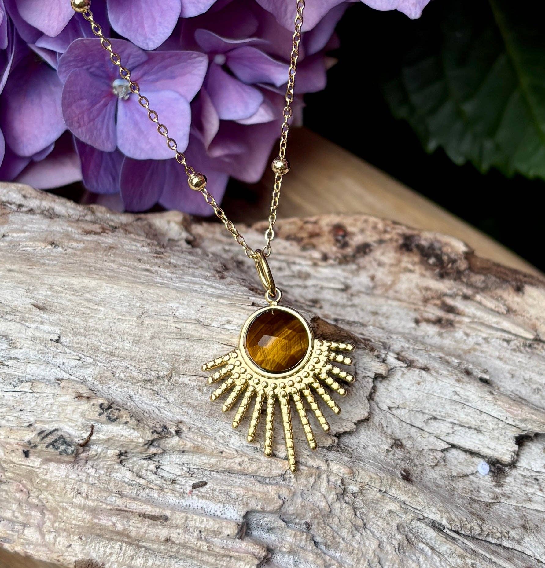 Gold necklace with a sun-shaped pendant on a wooden surface with purple flowers in the background
