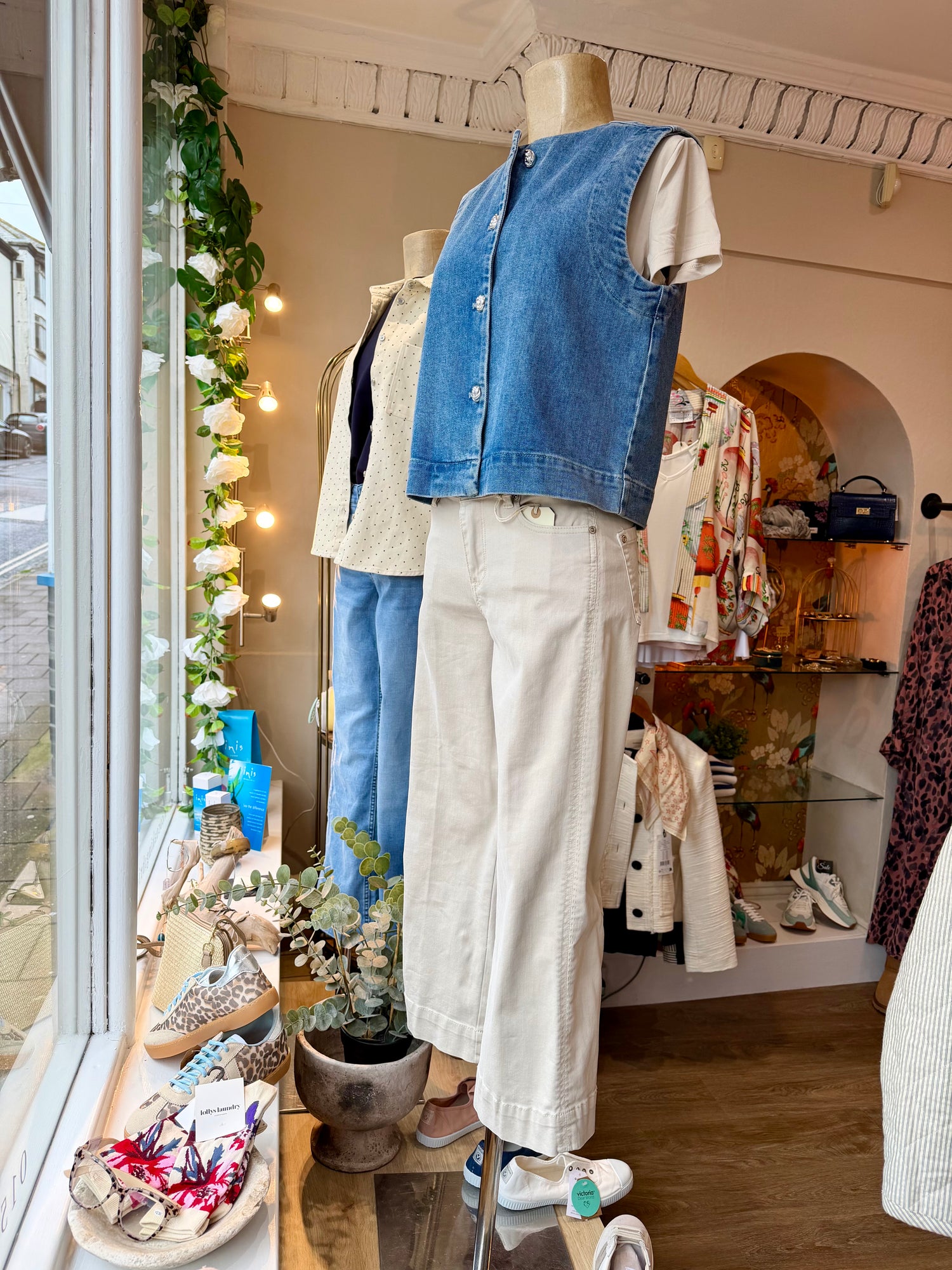 Mannequin wearing a denim vest and white pants in a store window with floral decorations.