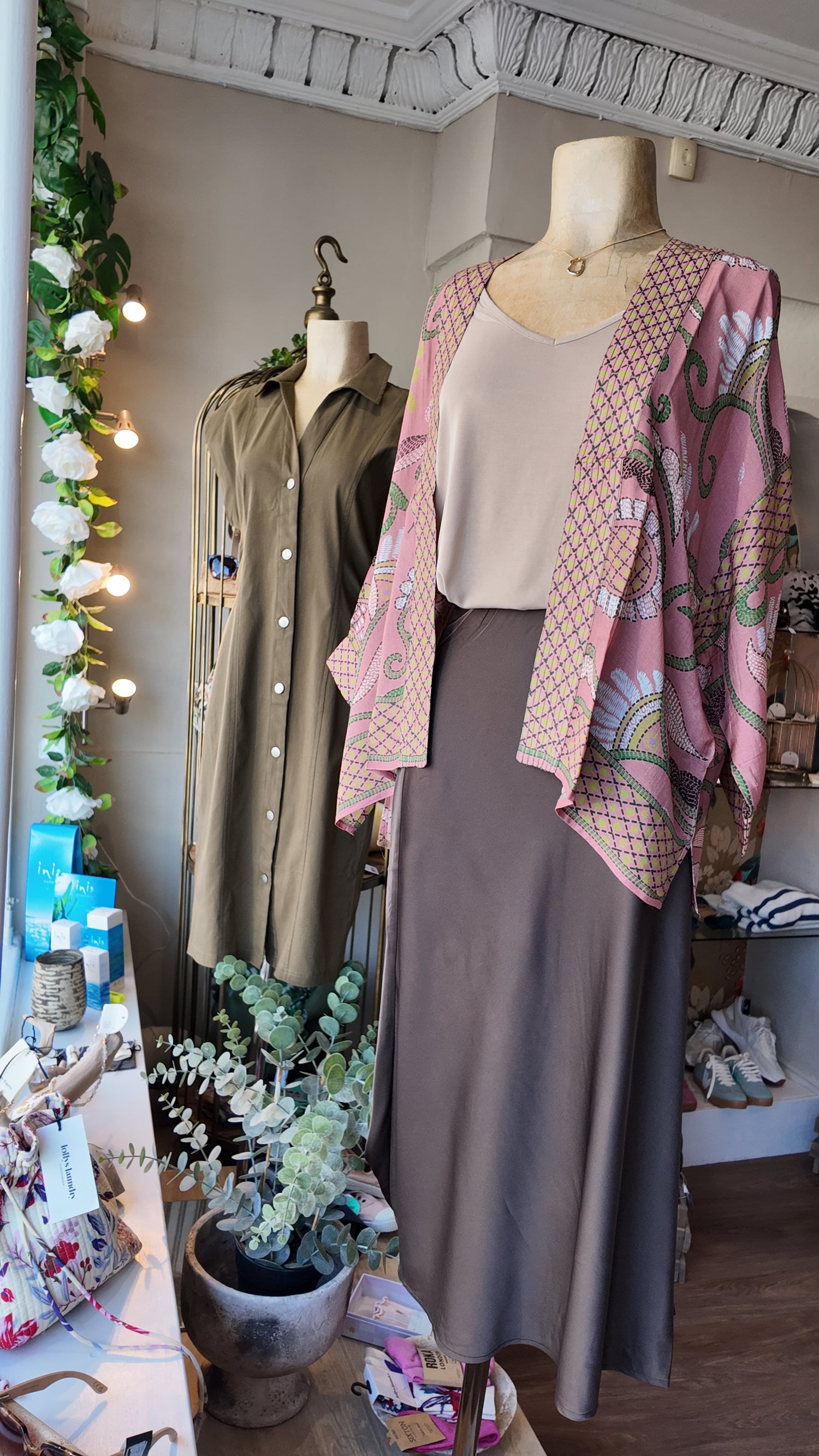A mannequin in a boutique displays a three-piece outfit consisting of a beige scoop-neck top, a brown midi skirt, and a pink patterned open-front cardigan. Another mannequin in the background wears an olive green button-up shirt.