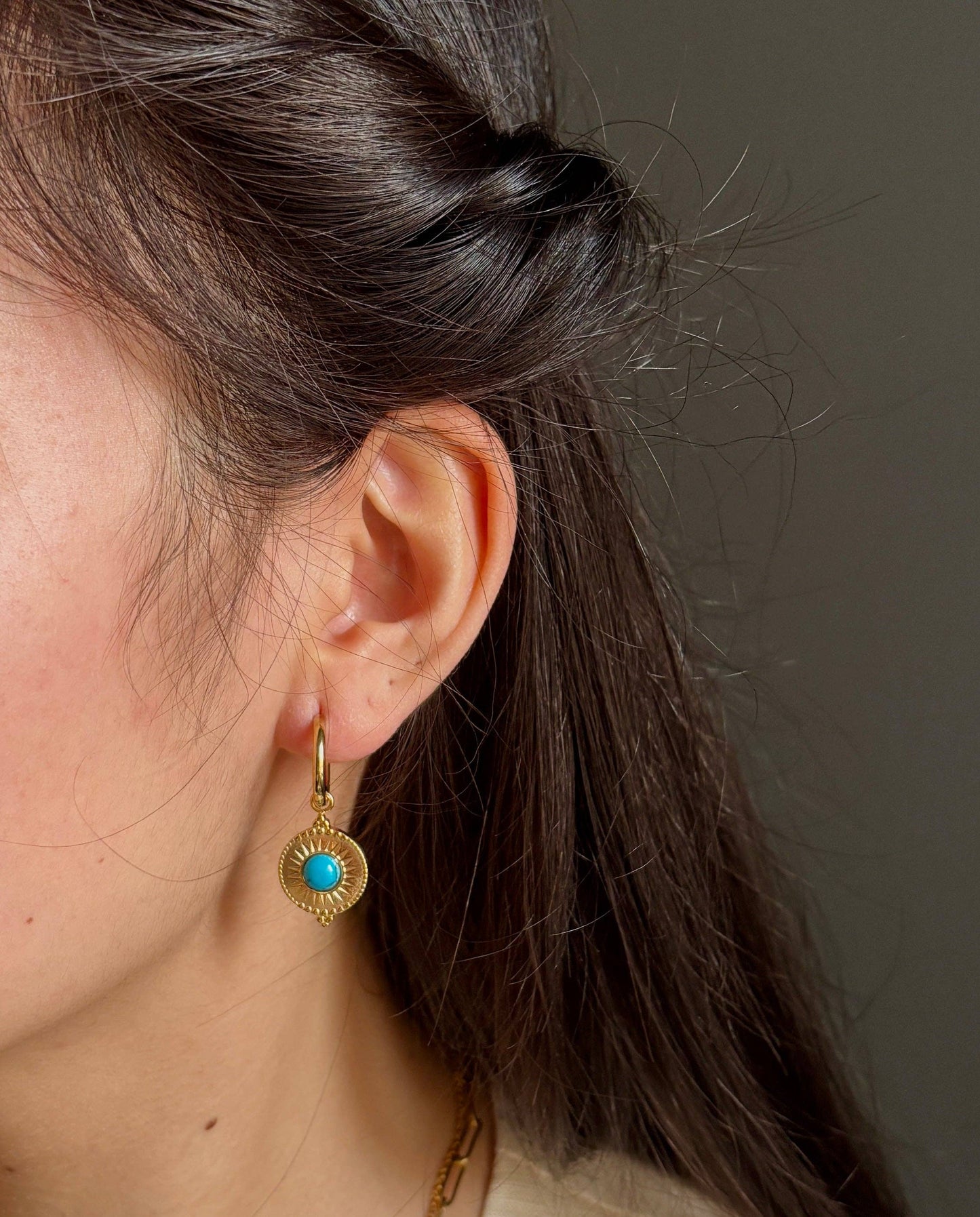Close-up of a person wearing gold earrings with a turquoise stone against a neutral background