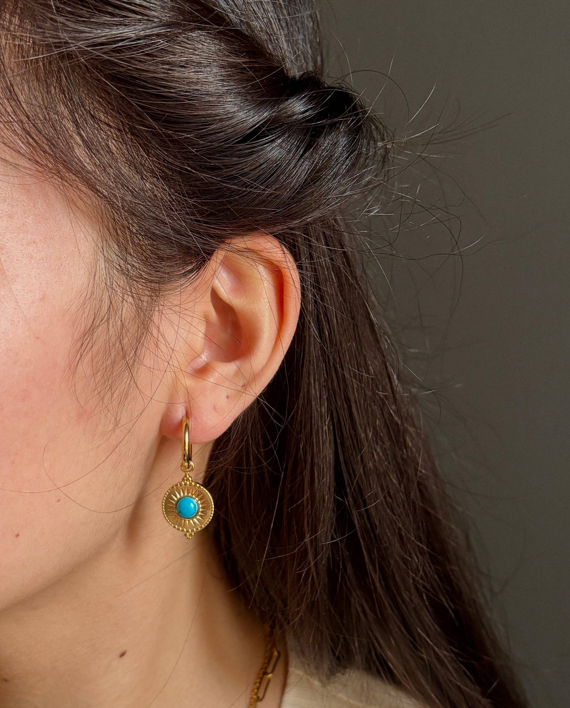 Close-up of a person wearing gold earrings with a turquoise stone against a neutral background