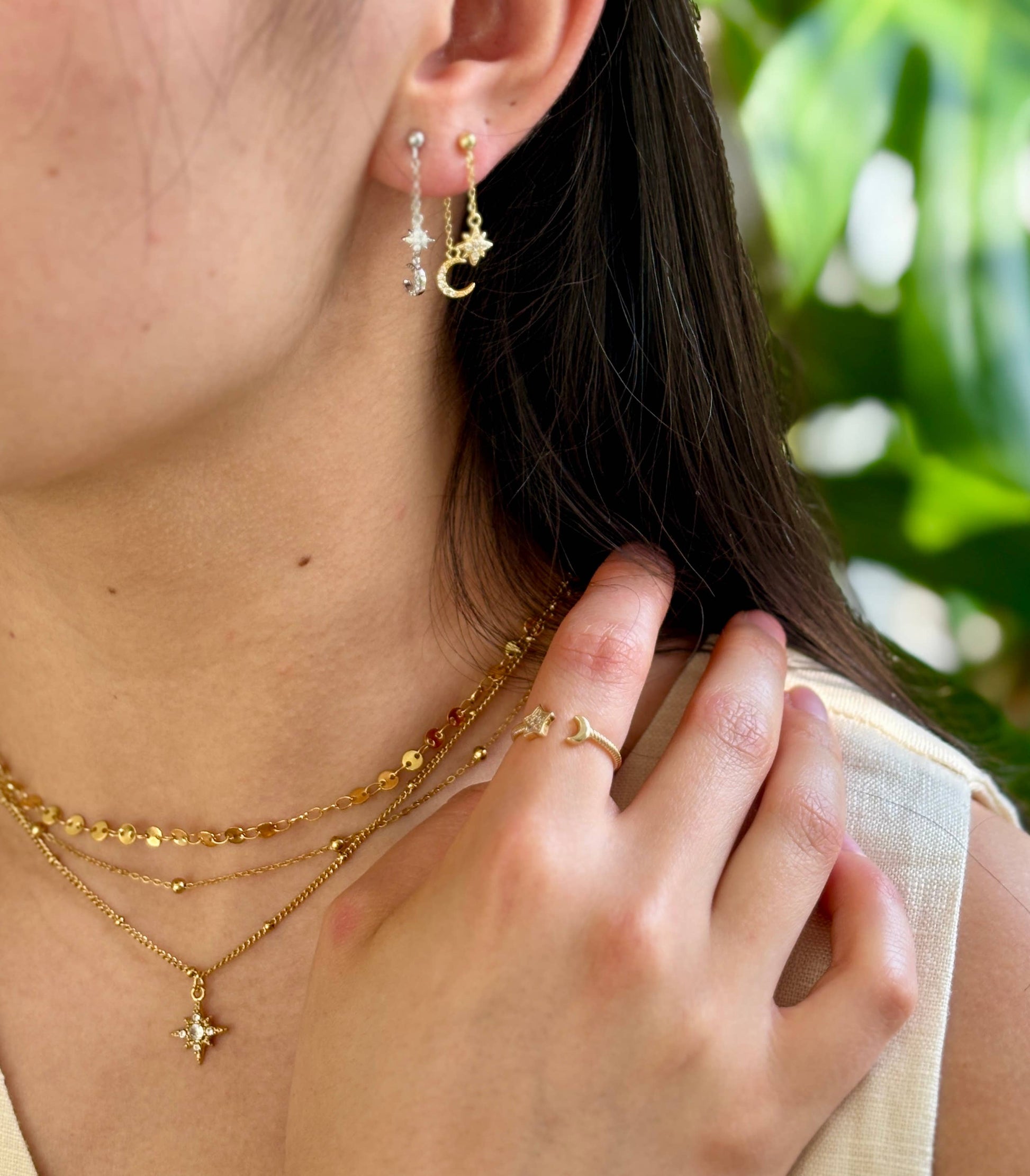 Close-up of a person wearing gold jewelry including earrings, necklace, and ring with a blurred green background.