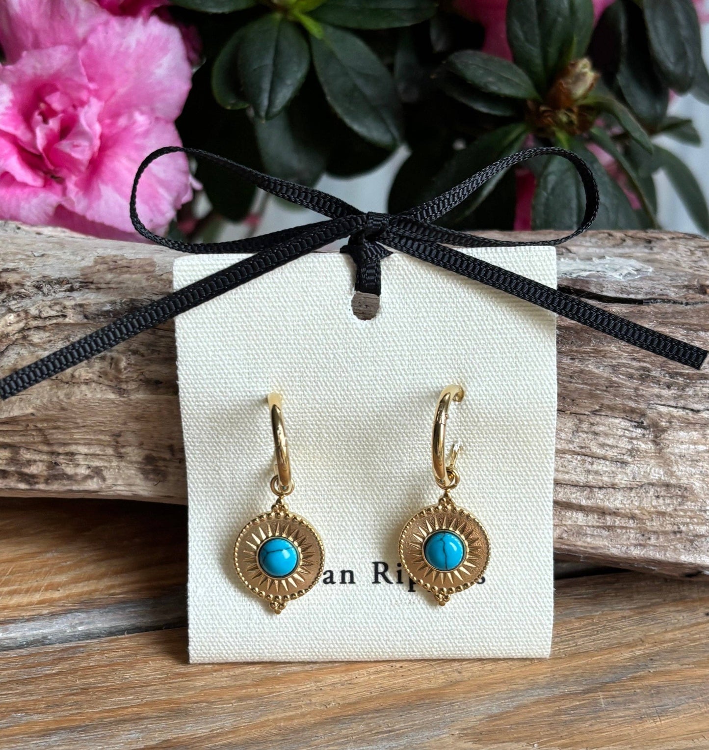 Gold earrings with turquoise stones on a white card with black ribbon, placed on wooden surface with pink flowers in the background.